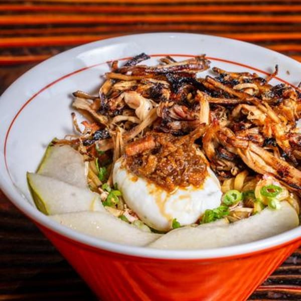 A vibrant bowl of noodles with shredded meat, a poached egg, and sliced green vegetables.