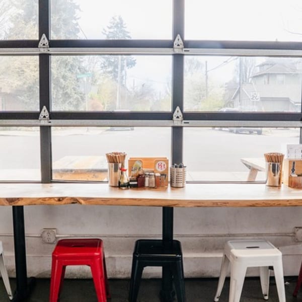 A long wooden counter with stools, condiments, and chopsticks, facing a large window overlooking a street.