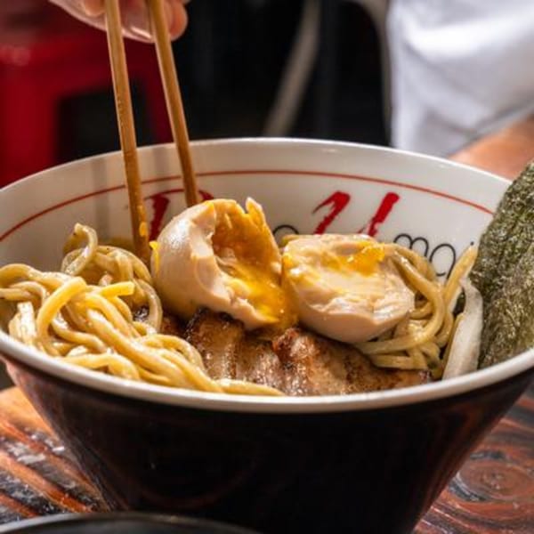 Chopsticks picking up noodles from a bowl of ramen with pork, soft-boiled eggs, and seaweed.