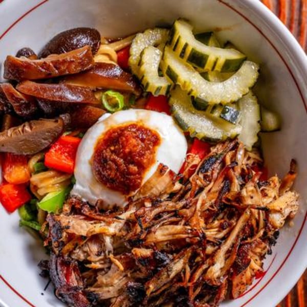A close-up of a colorful bowl with pulled pork, pickled cucumbers, mushrooms, and a soft-boiled egg.