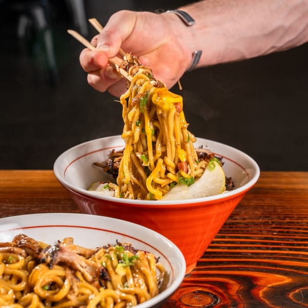 A hand with chopsticks lifts Baka Umai ramen noodles from a red bowl on a wooden table.