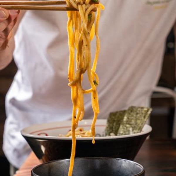 Chopsticks lifting long, saucy noodles from a black bowl, with a side of nori.