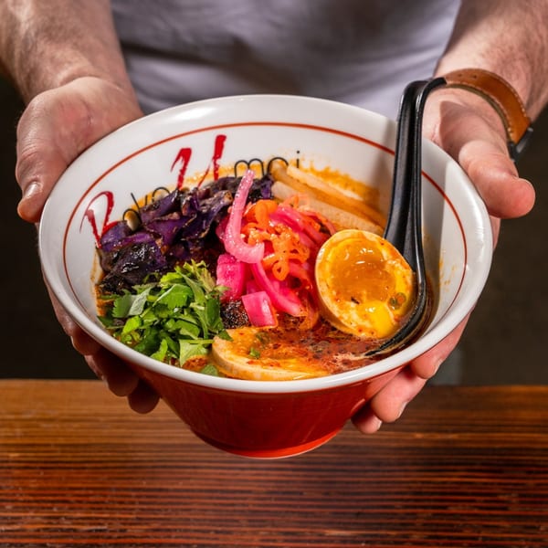 Hands holding a bowl of Baka Umai ramen with egg, pickled onions, and cilantro.