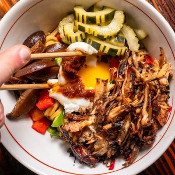 Chopsticks picking up a piece of mushroom from a bowl of food with egg, meat, and vegetables.