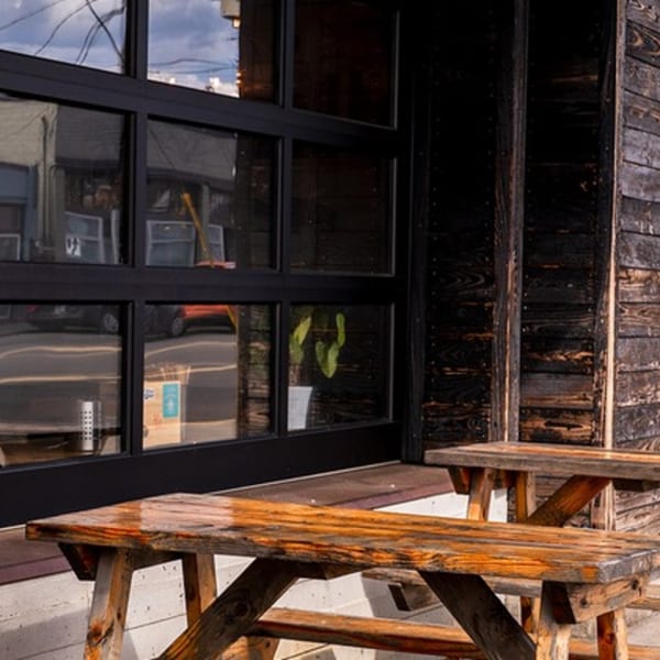 Two rustic wooden picnic tables sit outside Baka Umai, next to a large window and dark wood siding.