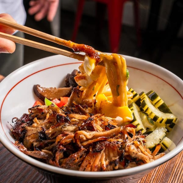 Chopsticks lifting cheesy ramen noodles from a bowl with grilled pork, mushrooms, and pickled cucumbers.